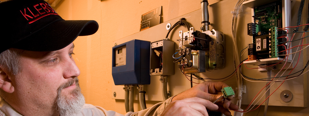 A KLEBS technician in a company cap inspecting and servicing a home heating system's controls and wiring.